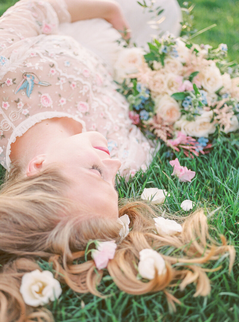 Blonde woman lays on grass with roses in her hair holding her baby bump, photographed by Maryland Maternity Photographer Marie Elizabeth Photography.