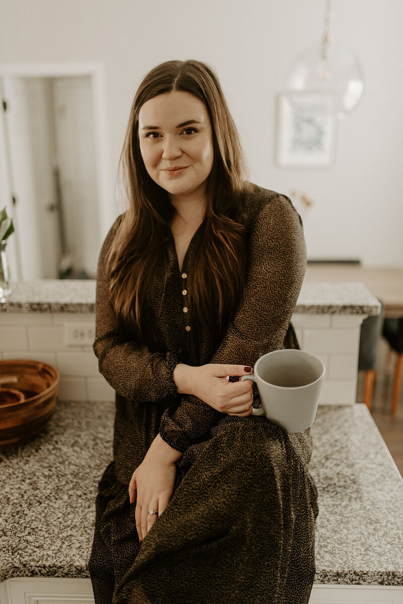 Copywriter sitting on the counter holding a blue coffee mug, smiling at the camera