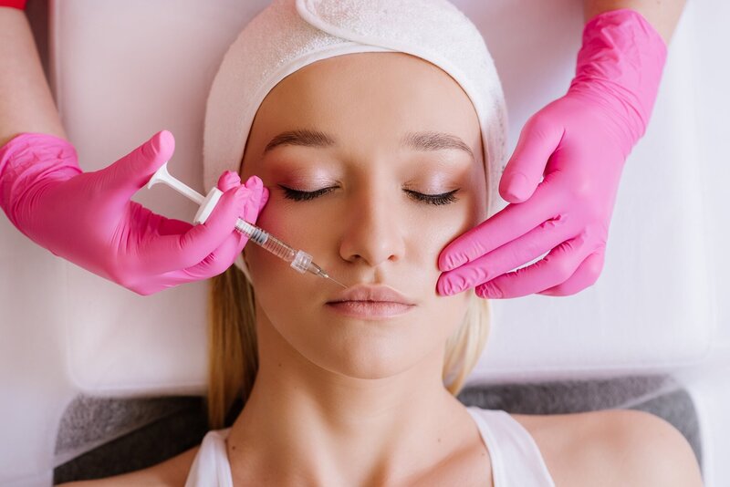 Closeup image of a woman lying down receiving a dermal filler injection from an esthetician wearing bright pink gloves