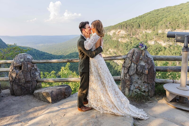 A bride and groom are embracing standing at a mountain overlook. They just says "I do" and are sharing a smile before their first kiss. The groom is holding the brides waist and the bride is holding the grooms face. The groom is wearing an olive green suit and the bride is in a long white lace dress.