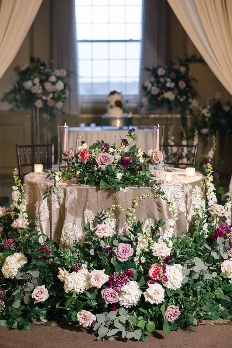 An elegantly decorated wedding table with a lace tablecloth is surrounded by lush floral arrangements of pink, purple, and white flowers. Candles are placed on the table, and a wedding cake is visible in the background under soft lighting.