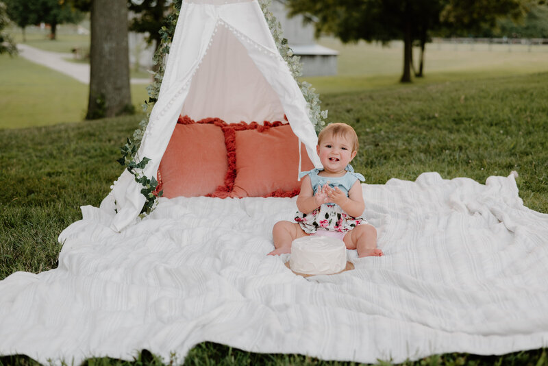 One year old girl sitting on white blanket at a farm in the grass with cake and white teepee behind her.