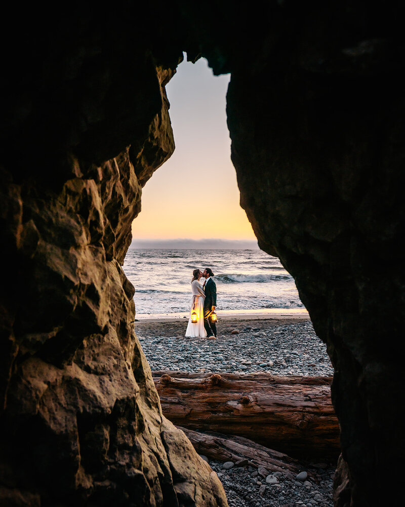 a couple in their wedding attire kiss in front of the ocean as they hold lanterns while the sun sets. Their adventure elopement photographers shoot through a hole in a large sea stack that frames the couple beautifully. 