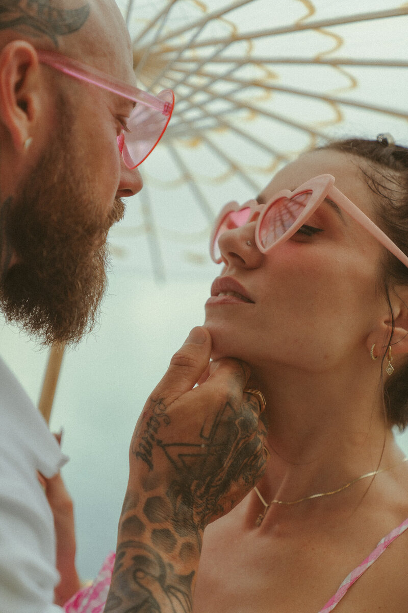 Close-up of tattooed couple wearing pink sunglasses sharing an intimate moment under a decorative parasol