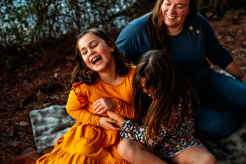 Two sisters and a mom are sitting on a fuzzy blanket with bushes behind them. The two girls are tickling each other and laughing hard. The mom is sitting behind them smiling at the girls.