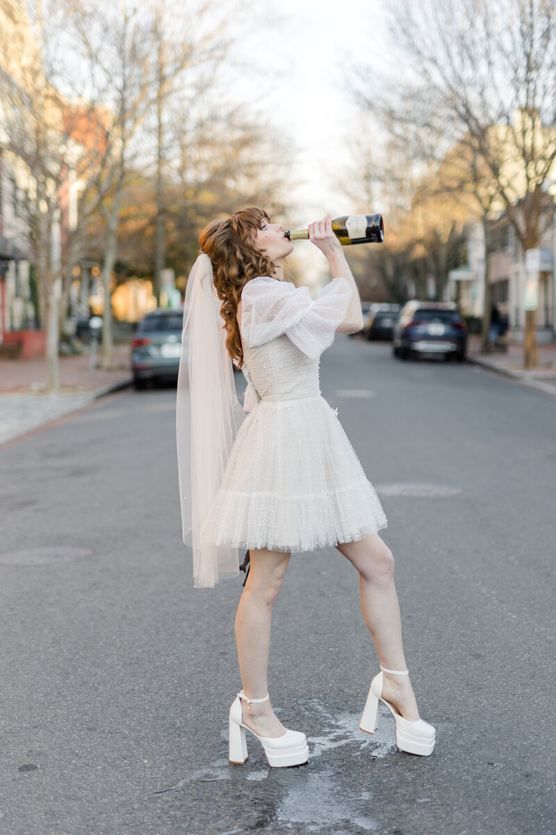 Queer bride in mini tulle sheer puff sleeve dress drinking champagne from the bottle standing in the middle of the street in Georgetown DC 