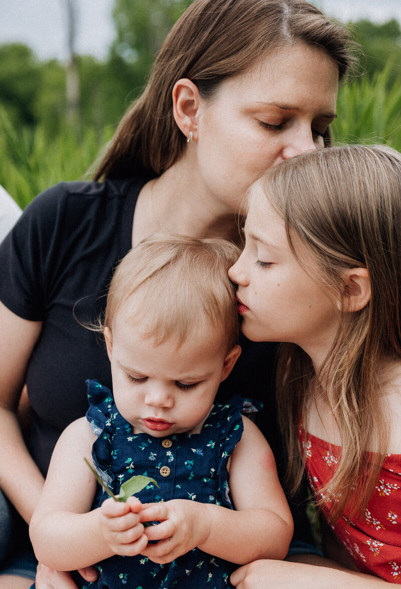 Close up image of mother kissing her oldest daughter and the daughter kissing her little sister while they sit in a field during a lifestyle family session. 