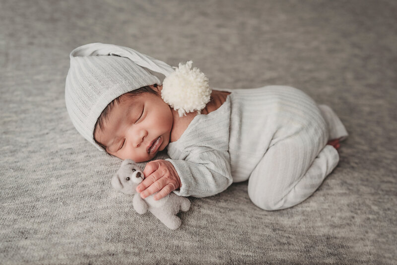 newborn baby boy holding teddy bear laying on belly in grey outfit grey hat and gray backdrop