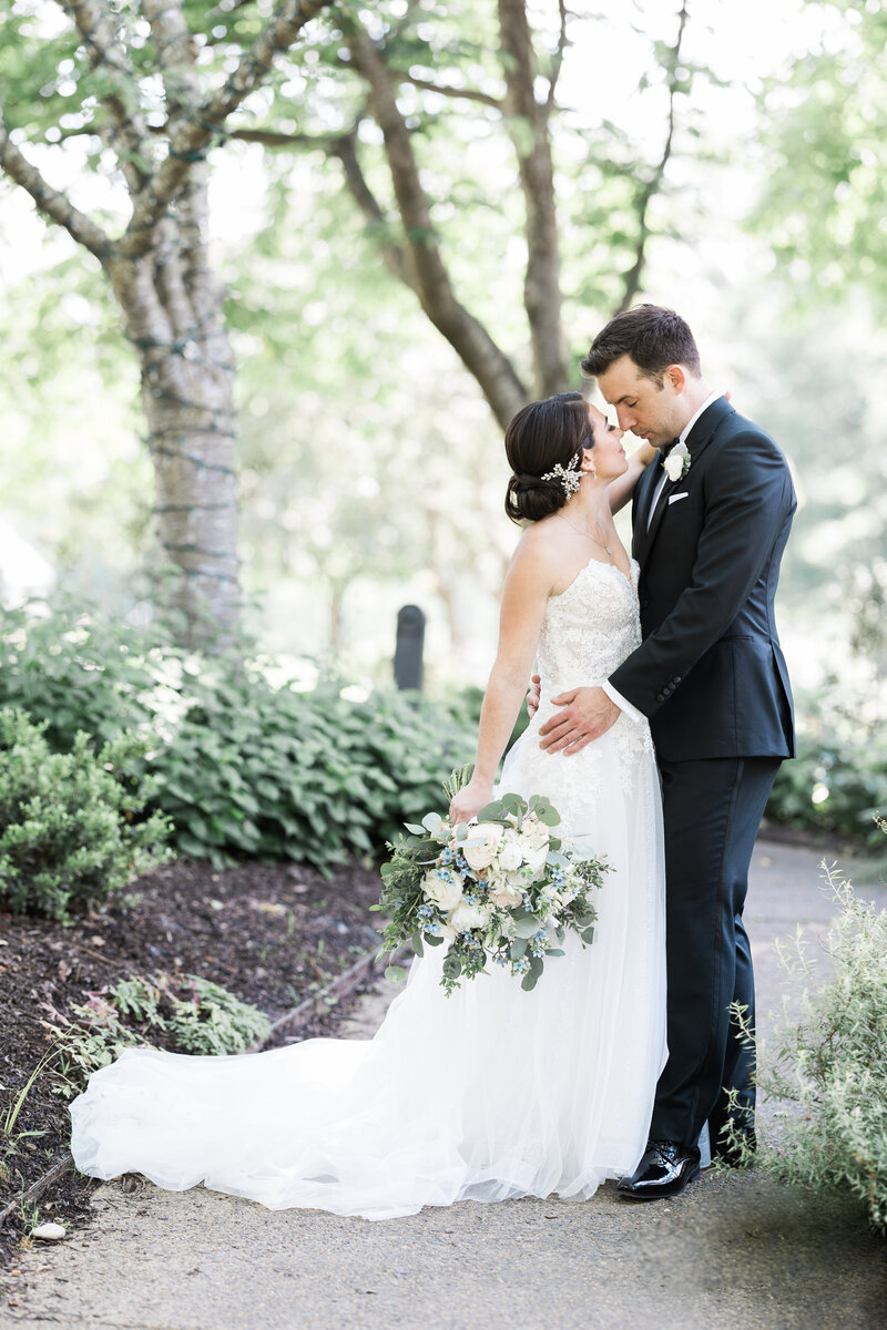 a white groom in a black tux facing and holding a white brunette bride in a strapless white gown with her white and blush bouquet at her side leaning into one another for an almost kiss