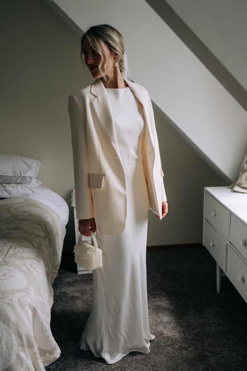 bride in ivory dress with blazer and pearl clutch in a bedroom by a window