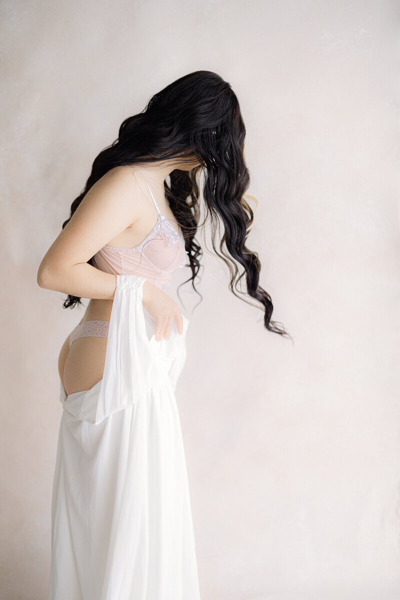 Woman with long dark brown curly hair puts on a white dress with her lingerie showing and her hair falling in front of her face during her Indianapolis boudoir photography session.