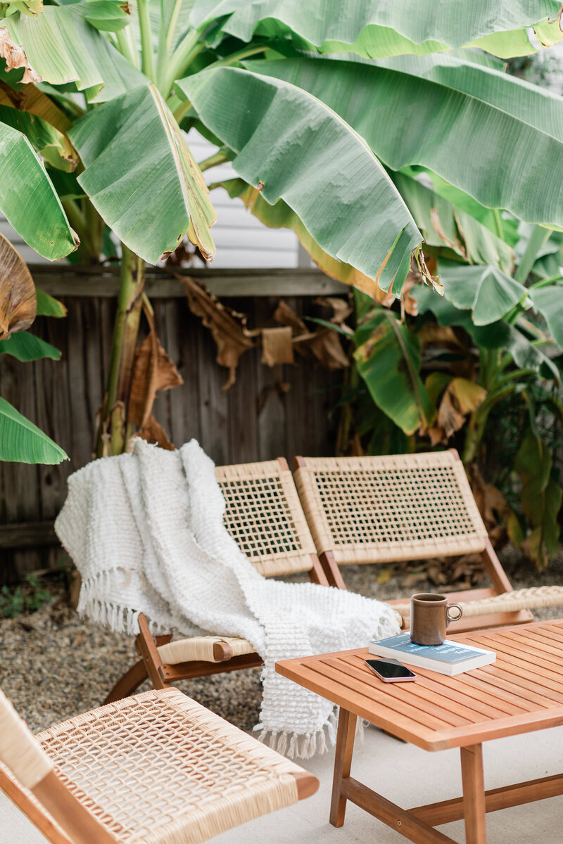 Outdoor San Diego backyard seating area with tropical plants, cane chairs, and a cozy throw blanket — lifestyle imagery for Mary Pernicano Real Estate.