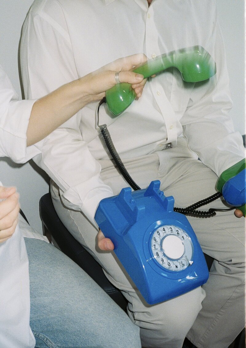 Two people in white shirts hold a bright green and bright blue rotary dial phones