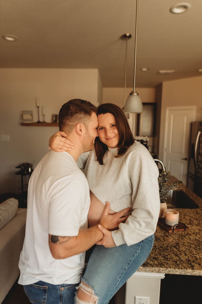 pregnant mother sits on the kitchen counter and lovingly wraps her arms around her husband