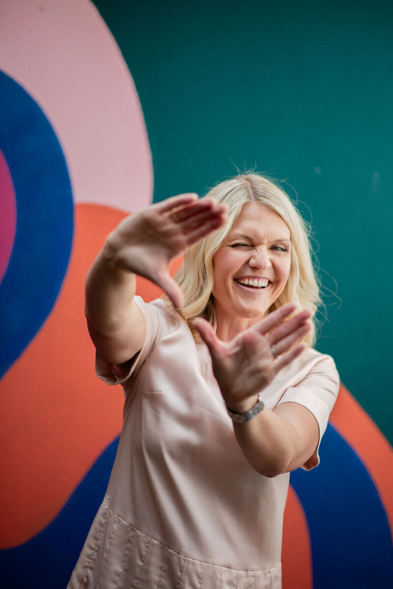 Emily Collins, business coach, smiling and framing her face with her hands in front of a colorful abstract mural