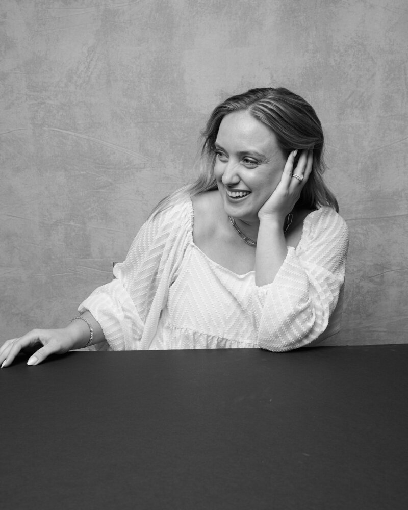 Black and white portrait of a woman in a textured white blouse, leaning on a table with a bright, joyful smile. Her hand rests gently against her face as she laughs, exuding warmth and confidence. A candid, unfiltered moment from 'THIS IS ME – A Photography Experience by Katie Campbell' in Atlanta, GA, celebrating authenticity and self-expression.