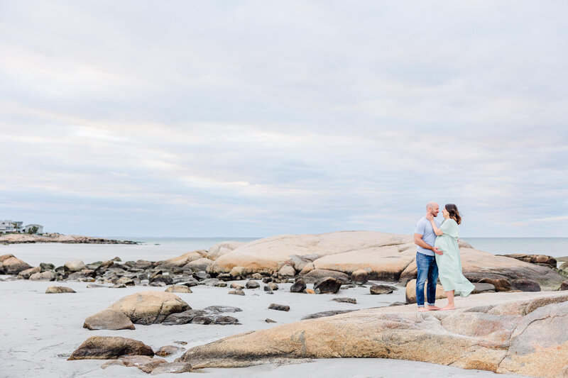 Pregnant woman and her husband stand close on a rock at a beach during a Boston Maternity Photography Session