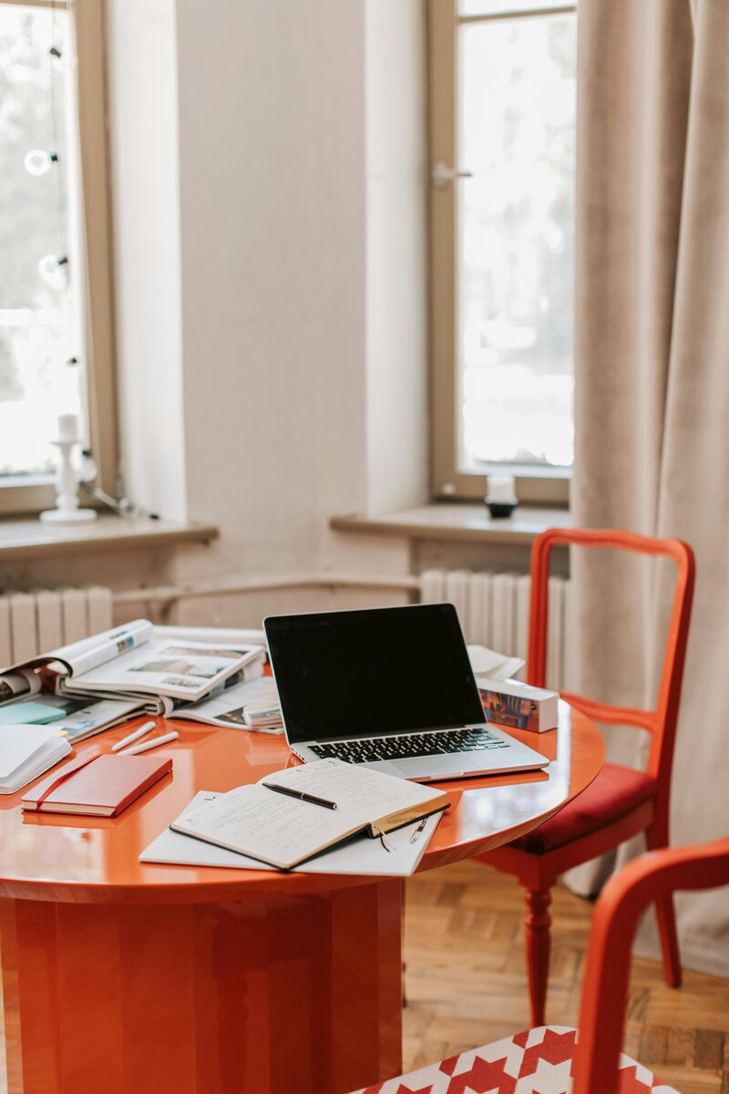 Laptop, notebooks, and papers spread out on a red table in a bright room with orange chairs and large windows.