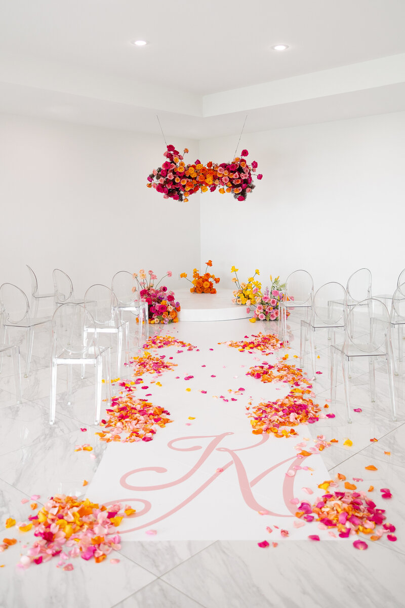 Acrylic Queen Chairs in an all white room, practically invisible with bright orange and red and pink flower petal aisle to ceremony pedestal.