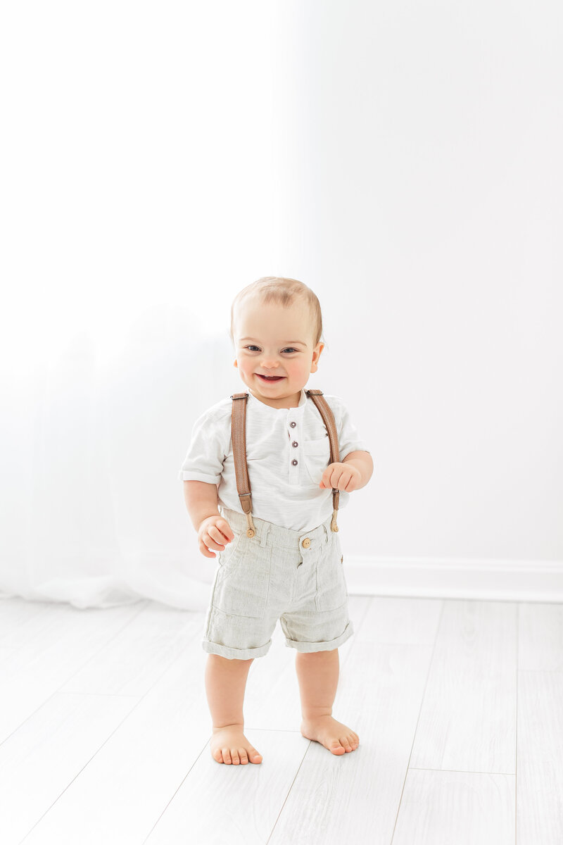 A northern va family photographer photo of a happy baby boy wearing suspender shorts and a white henley shirt standing in front of a window