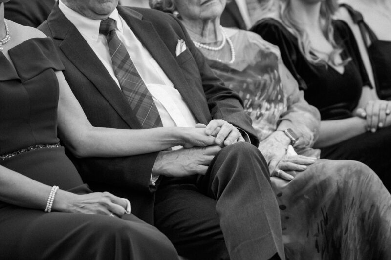 A close-up of two hands clasped together, surrounded by seated family members dressed elegantly, conveying intimacy and support for newlyweds
