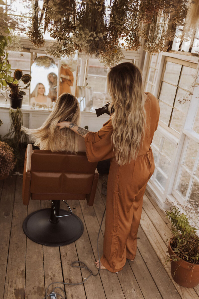 The hair extension specialist is working with her blonde client in Dallas, Texas. Behind them the salon has small windows and is decorated with green leaves on the ceiling.