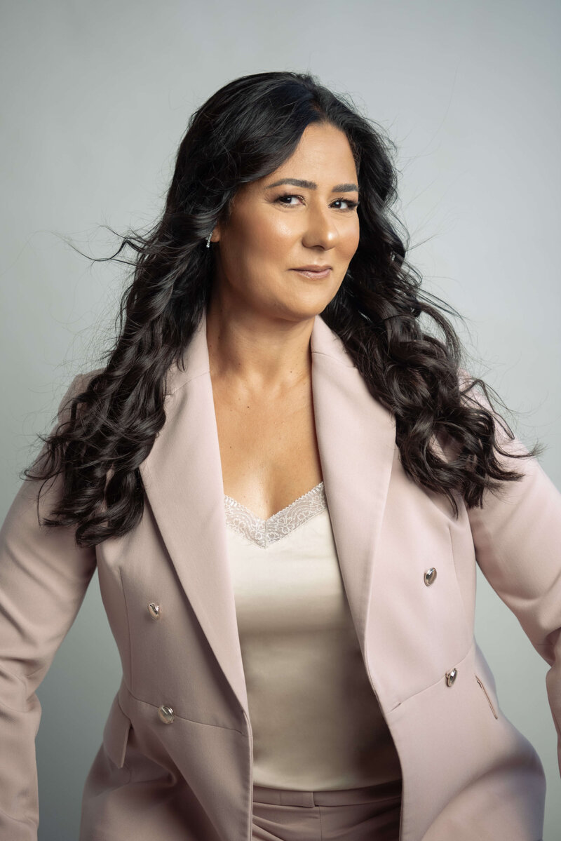 Portrait of Asmahan seated on wooden stairs, wearing a white top and black pants, with a calm and confident expression in a moody, editorial setting