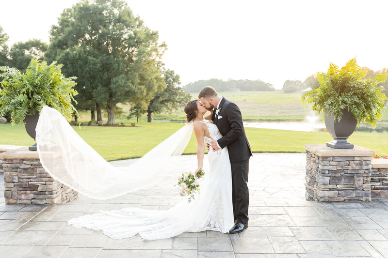 A newlywed couple shares a romantic kiss at sunset, with the bride’s veil billowing in the wind, set against a picturesque countryside estate