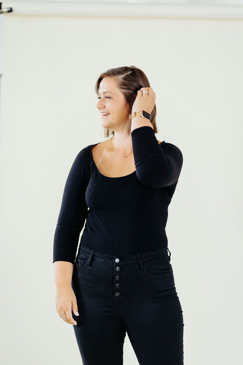 Kira from Kira Writes Copywriting posing in front of a tan background wearing all black while looking to the left and swiping hair back.