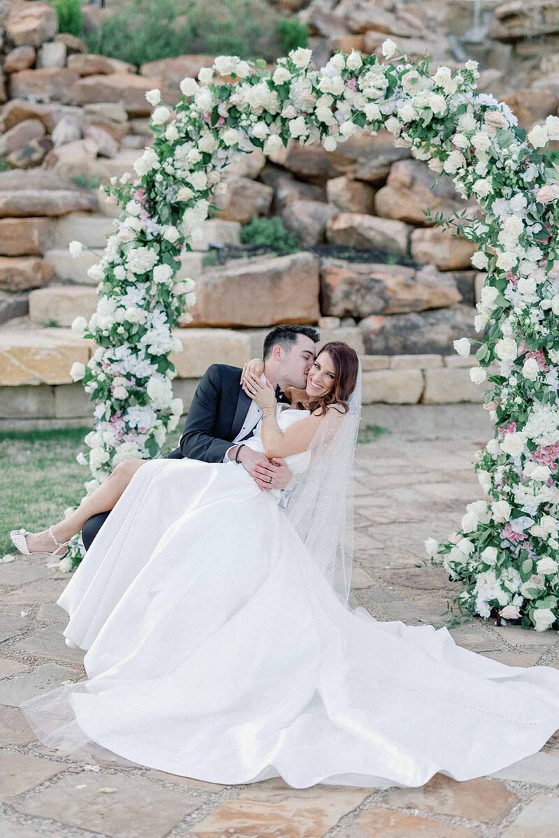 groom kisses bride in front of luxury floral arch