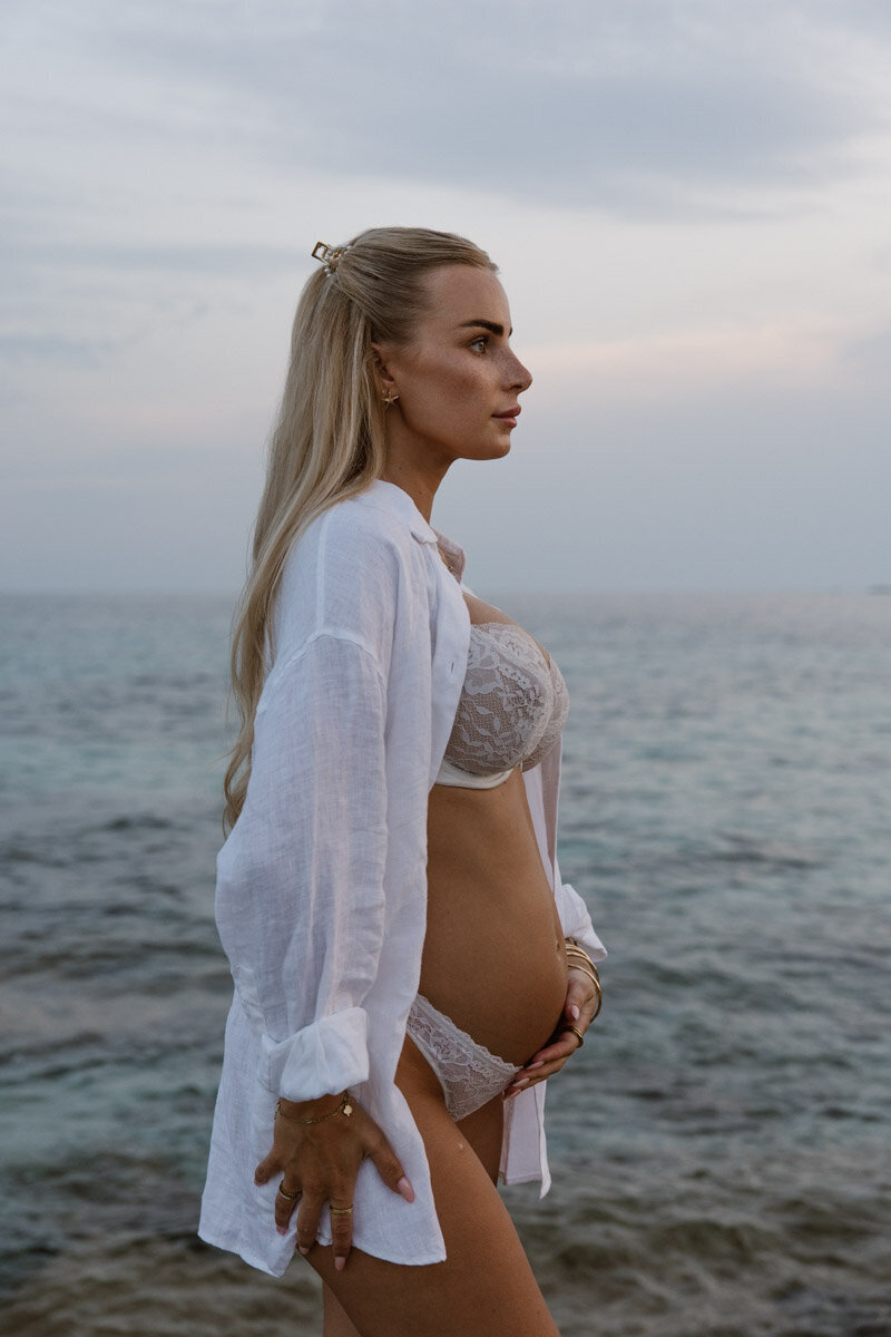 A pregnant woman in lingerie is posing in front of the sea, holding her belly.