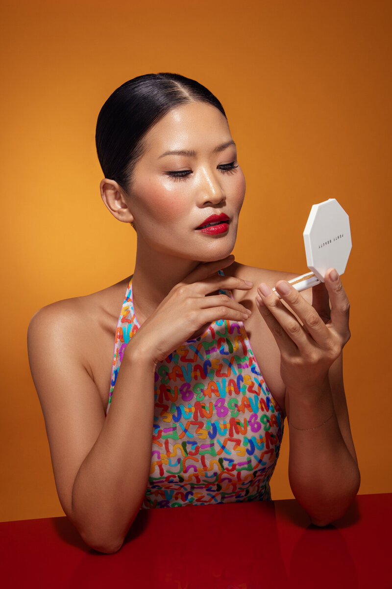 Danielle Yu applying lipstick while looking into a compact mirror, captured against a vibrant orange background by Ylva Erevall.