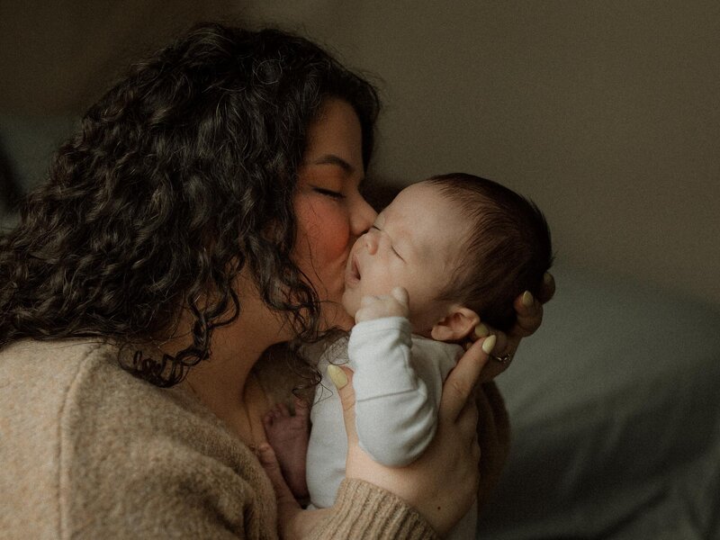 Mother holding and kissing her newborn baby against a soft neutral background, capturing a tender, intimate moment.