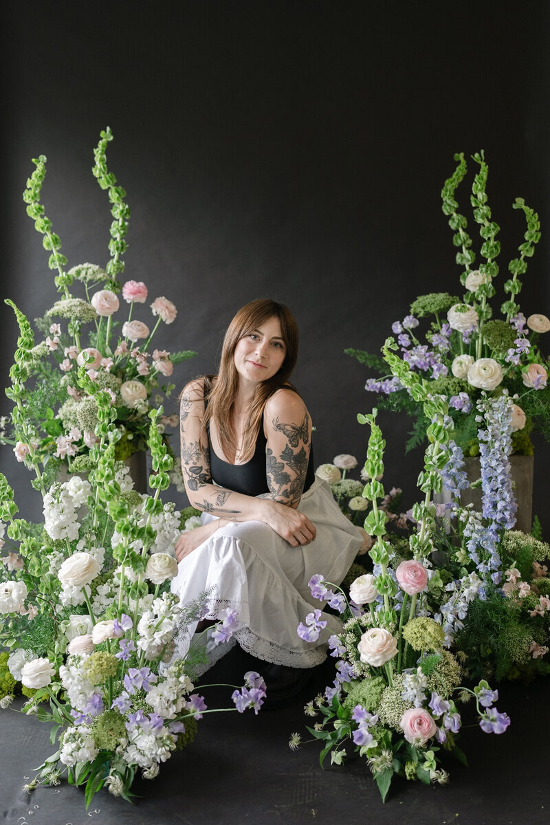 A florist posing among purple, green and white floral arrangements against a black backdrop. 