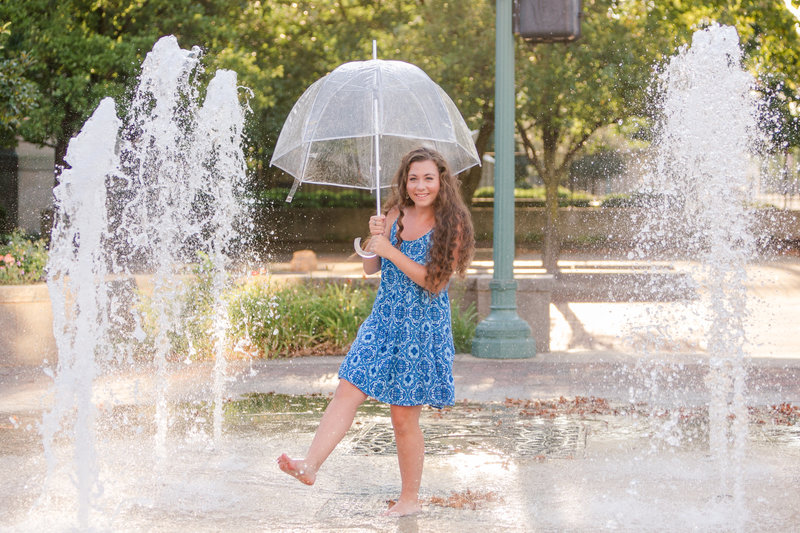 perry high school senior girl, in blue summer short sundress holding a clear umbrella kicking water in fountains downtown canton ohio photographed by jamie lynette photography canton senior photographer