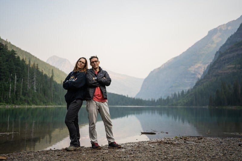 saint augustine photographers rob and jill stand in front of a lake with mountains in the background from an elopement session that they photographed in Glacier National Park