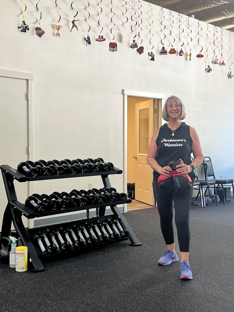 Coach Lori of the Oregon Parkinson's Warriors Foundation smiles and holds boxing gloves next to a stack of weights inside the boxing gym.
