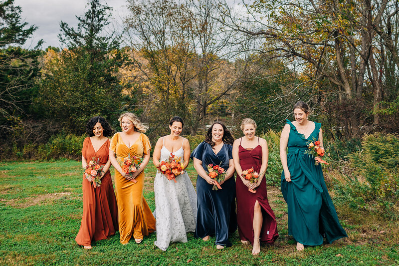A bride and her wedding party wearing shades of terra cotta mustard navy teal and maroon and carrying mixed seasonal bouquets