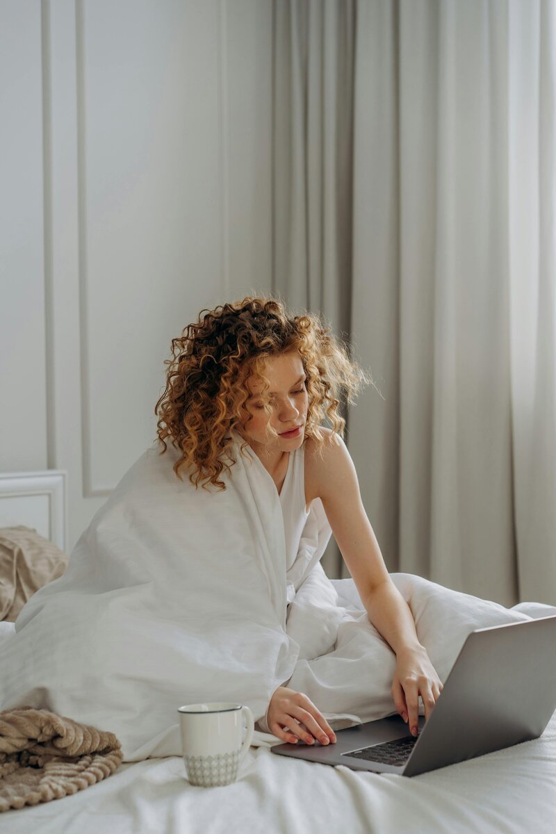 Woman working on a laptop from bed with a coffee cup, focused on client support tasks