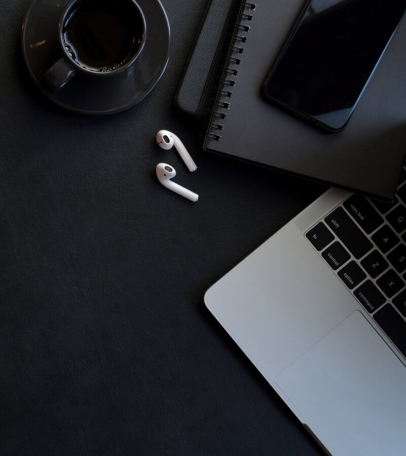 A workspace setup with a black coffee cup, a pair of white wireless earbuds, a spiral notebook, a smartphone, and the corner of a laptop on a dark surface. The items are arranged neatly, giving a minimalist and organised appearance.