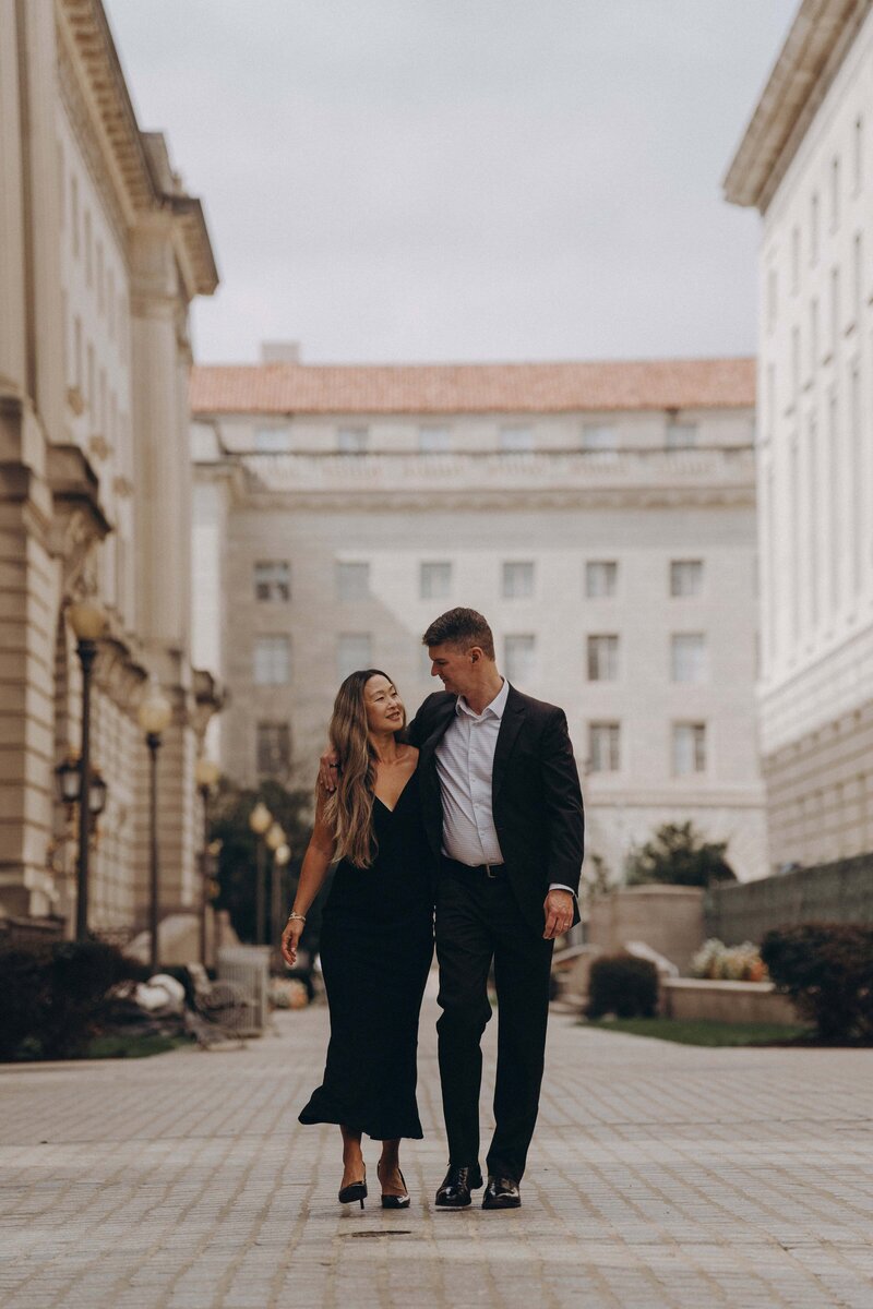A couple strolls arm in arm along a cobblestone path, surrounded by elegant architecture and a serene urban backdrop.