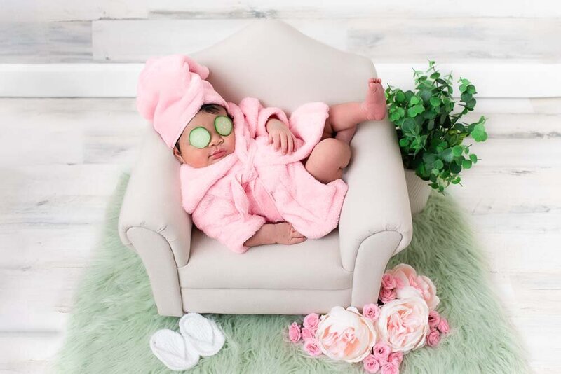 Newborn baby girl in a pink robe with cucumber slices on her eyes, posed on a miniature chair prop with flowers and greenery — spa-themed newborn photography by Misty Dawn Photography in Birmingham, AL.