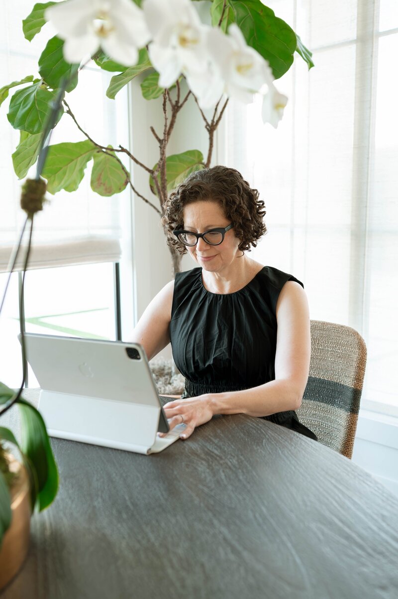 The owner of Scout & Bimble types on a tablet at a dining table near tall windows, with bright natural light and a large fiddle-leaf fig plant beside her.