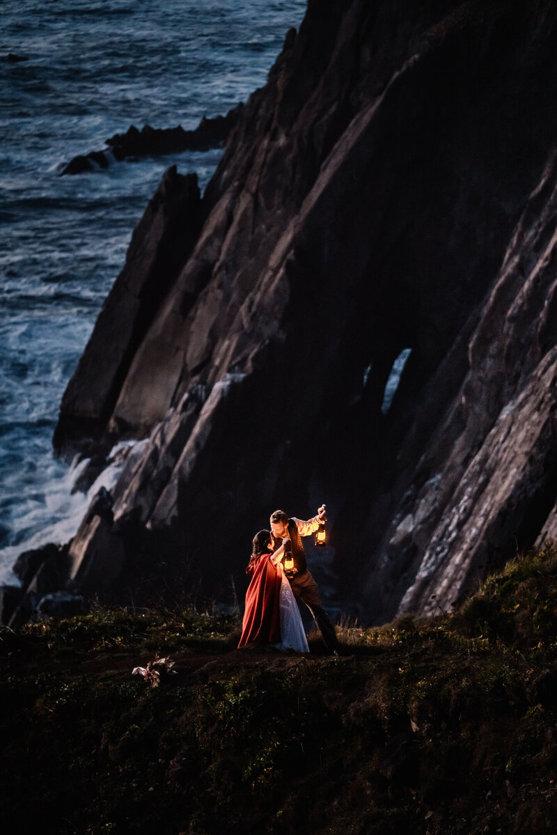 During their Oregon Coast elopement , a bride and groom in their wedding attire stans on a coastal cliff. They hold lanterns as they kiss during  blue-hour
