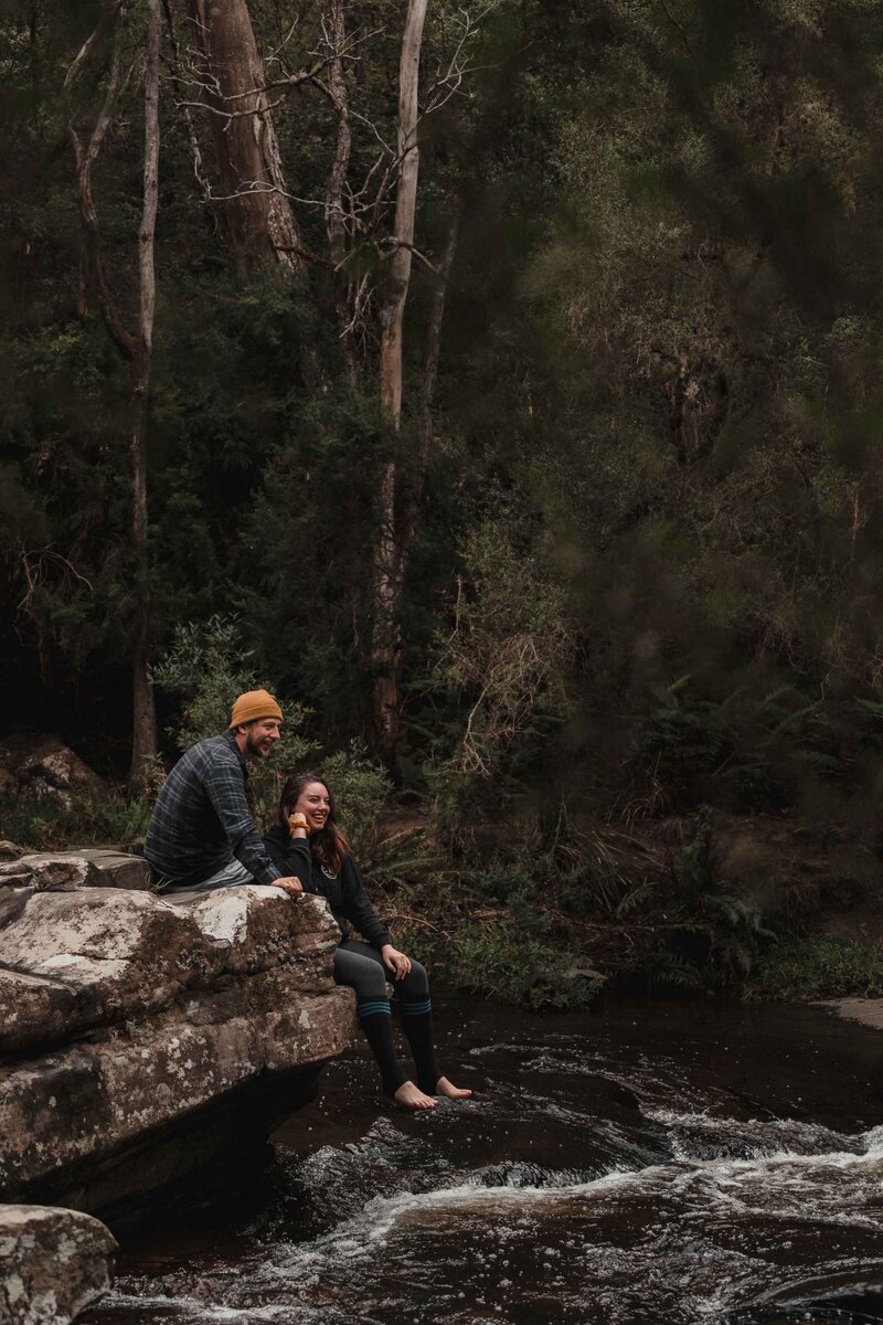 Couple sit on rock overhanging river. Peter Ali and his partner Emma