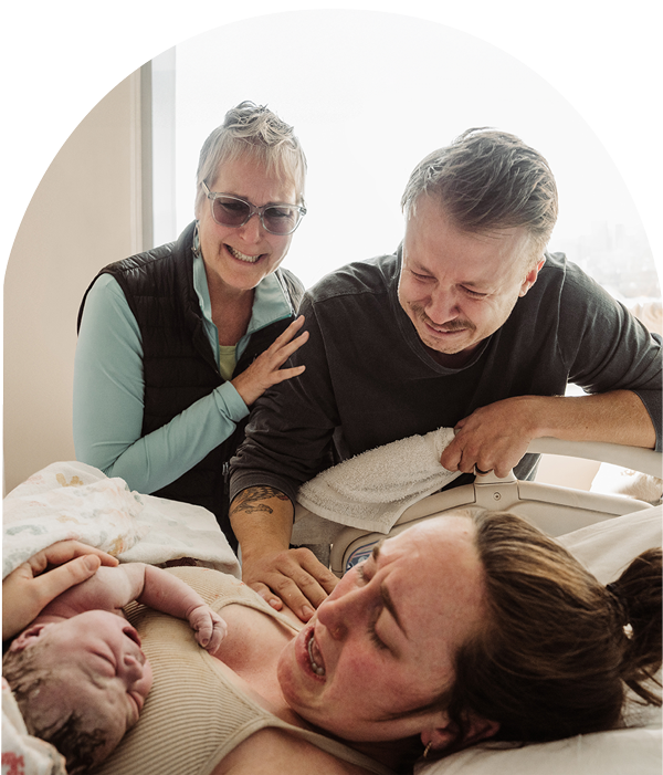 dad and grandmother cry looking down at a newborn baby held by her mother