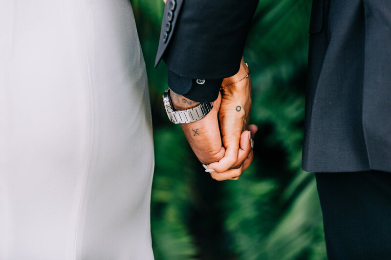 A close-up of a bride and groom holding hands. The bride is wearing a white gown, and the groom is dressed in a dark suit. The groom has tattoos on his hand and is wearing a silver wristwatch. They are standing in front of a green, blurred background.
