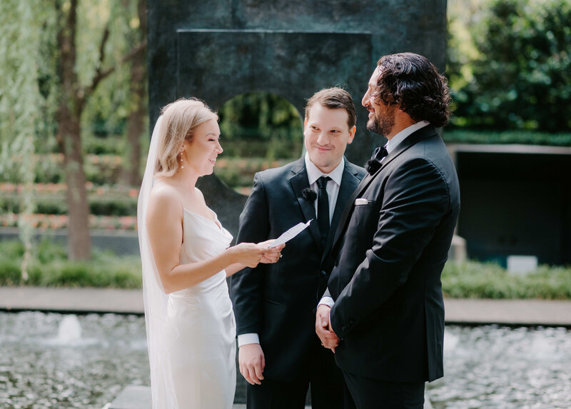 Outdoor wedding ceremony by a fountain with a bride reading vows to her groom, accompanied by an officiant in a black suit