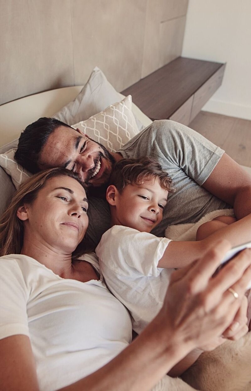 A smiling family of three rest in bed as the morning sun shines in. This could represent the strong bonds the family has after meeting with a marriage counselor in Florida. We offer infidelity recovery, couples therapy and marriage counseling, and other services. Contact a Florida therapist today for support.
