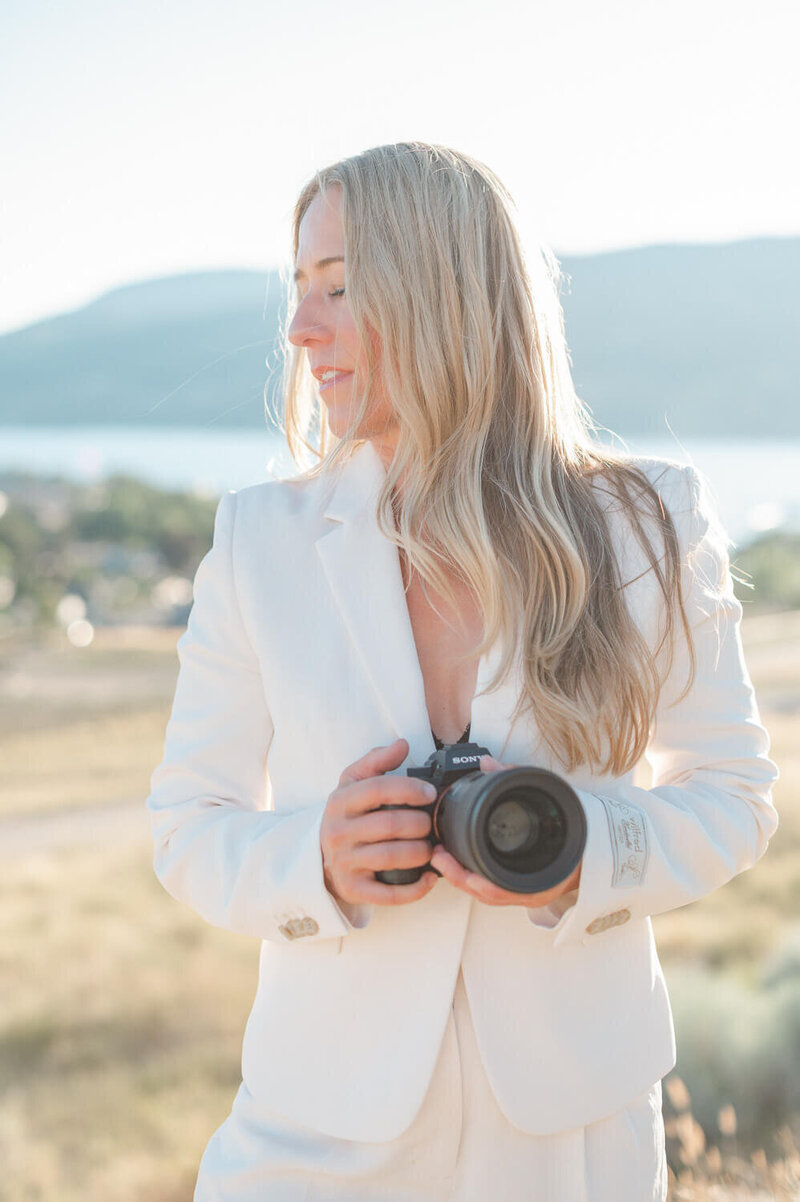 Kelowna brand photographer Kristen Clancy wearing white suit outdoors and holding camera looking off to the side